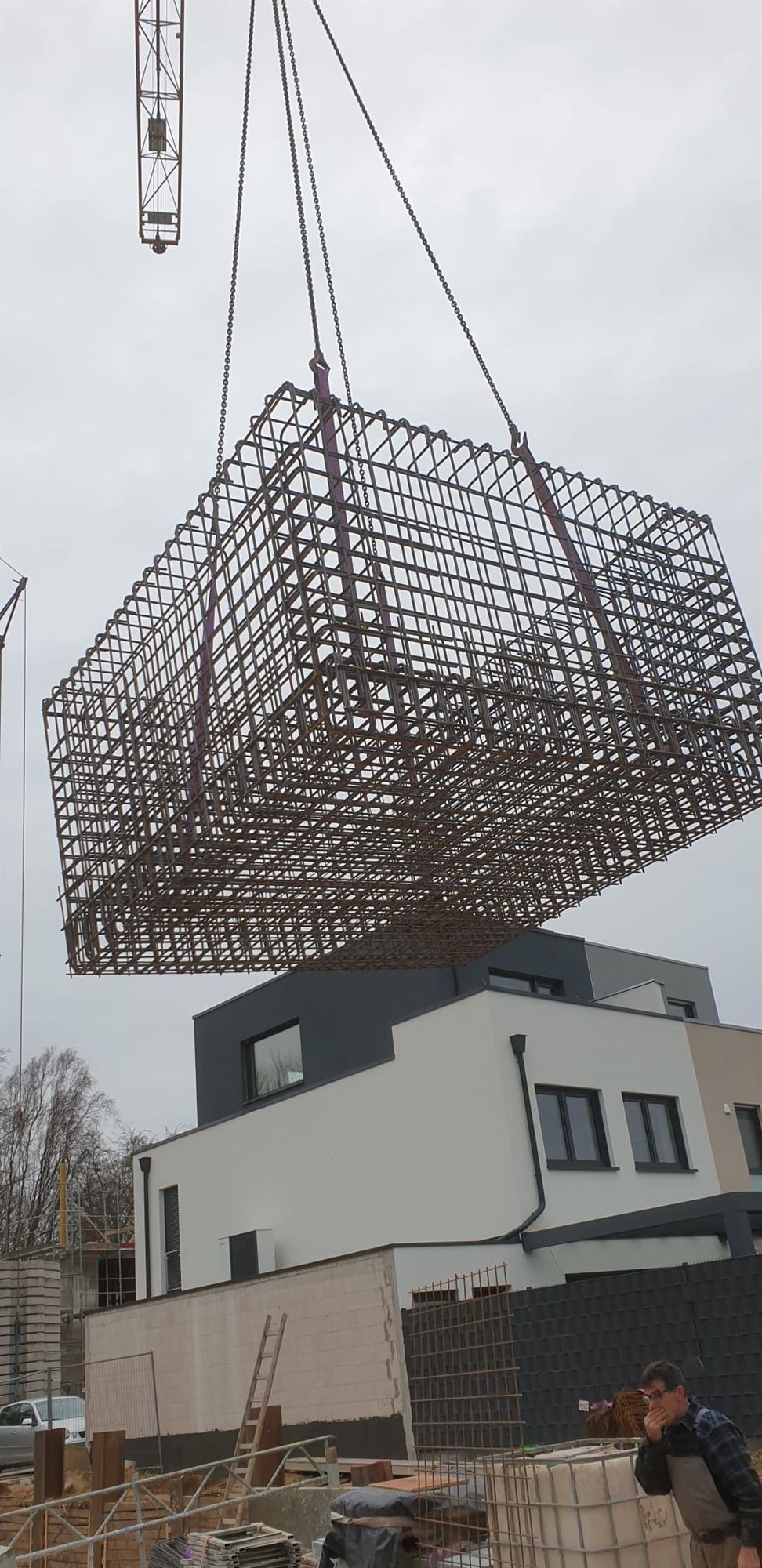 Bewehrungskorb aus Stahl über einer Baustelle, im Hintergrund ein fertiges Wohngebäude